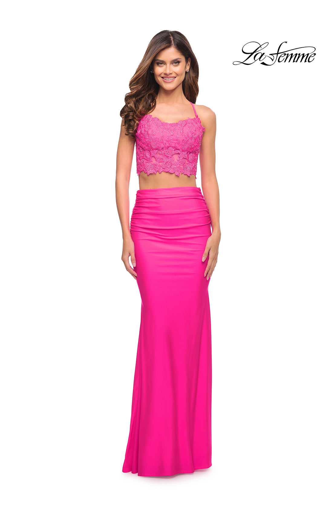 A woman smiles in front of a white background, wearing the Neon Pink Two-Piece Long La Femme Prom Dress with a lace crop top and fitted skirt.